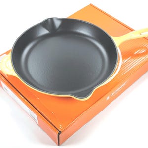 May include: A yellow cast iron skillet with a black interior sits on top of an orange box. The skillet has a long handle and a rounded shape. The box has the brand name printed on it. The skillet is new and unused.