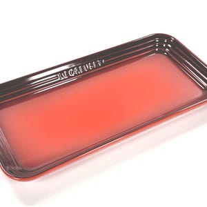 May include: A rectangular Le Creuset serving tray featuring a gradient finish that transitions from a vibrant orange center to a deep, dark red rim. The tray has raised, ridged edges and the brand name Le Creuset is embossed on one side.