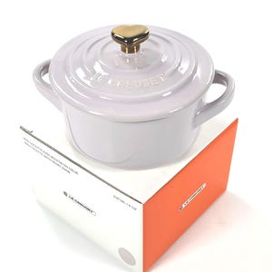 May include: A lavender-colored Le Creuset mini cocotte with a heart-shaped gold knob on the lid. The small, round pot has two handles and sits atop a white and orange box. The box has the Le Creuset logo and text.