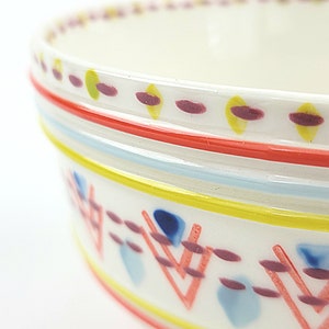 May include: A white ceramic bowl with a colourful geometric pattern around the rim. The pattern includes red, yellow, blue, and purple lines and shapes.
