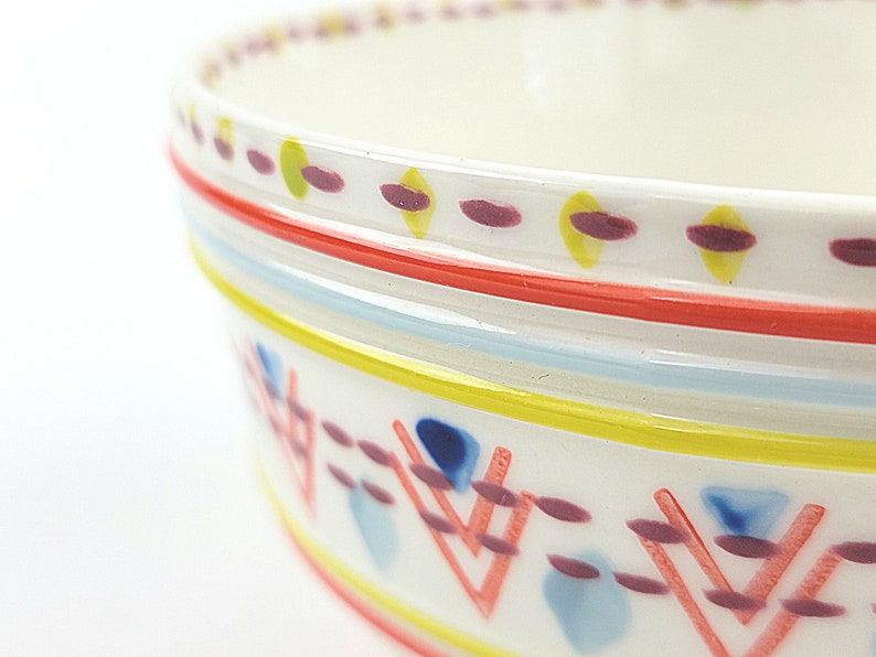 May include: A white ceramic bowl with a colourful geometric pattern around the rim. The pattern includes red, yellow, blue, and purple lines and shapes.