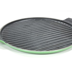 Op de afbeelding: Een ronde grillpan van gietijzer met een geribbelde kookoppervlak. De pan is zwart met een groene rand en handgrepen. De handgrepen zijn gebogen en geribbeld. Deze pan is ontworpen om te grillen, met een diameter van ongeveer 30 cm.