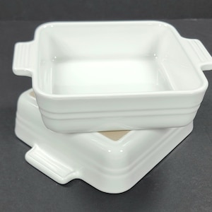 Peut inclure: Deux plats à four carrés en céramique blanche empilés. Chaque plat a une forme rectangulaire avec des coins arrondis et deux poignées. Les plats ont une finition brillante et sont posés sur un fond sombre. Idéal pour la cuisine.