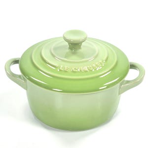 May include: A light green Le Creuset casserole dish with a matching lid and two side handles. The lid has a small knob and the brand name is embossed on the lid. The pot is made of enamelled cast iron.