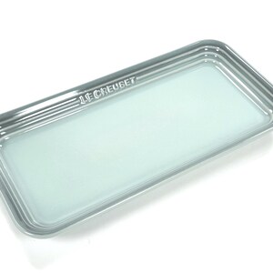 May include: A rectangular Le Creuset serving tray in a pale seafoam green color with a glossy finish. The tray features a raised rim with three concentric embossed lines and the brand name Le Creuset embossed on one of the shorter edges.