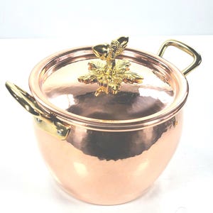 以下が含まれることがあります： 金色の花の模様が付いた蓋付きの銅製の調理鍋。鍋には2つのハンドルがあり、光沢のある反射面があります。蓋には、詳細な金の花の装飾が施されています。クラシックなキッチン用品です。