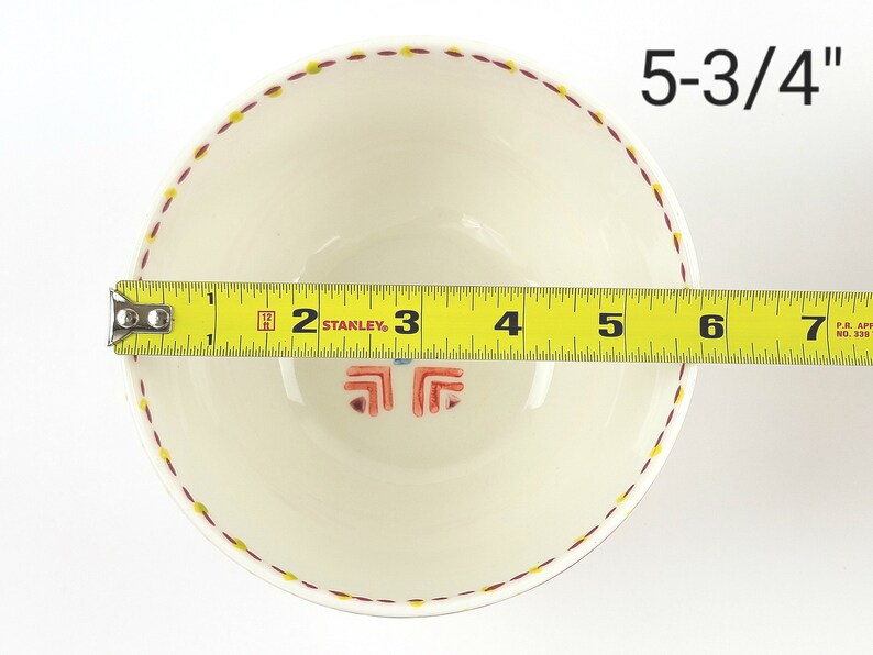 May include: A white ceramic bowl with a red and yellow patterned rim. The bowl measures 5-3/4 inches in diameter.