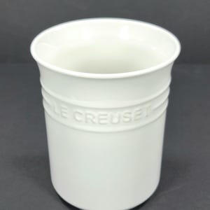 May include: A white Le Creuset utensil holder. The cylindrical holder has a wide opening and a slightly flared rim. The brand name is embossed around the middle of the holder. The holder is made of a solid, opaque material.