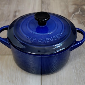 May include: A round, deep blue enameled cast iron Dutch oven with two side handles and a matching lid. The lid features a black knob and the embossed text LE CREUSET. The pot has a glossy finish and a circular design with concentric rings on the lid.