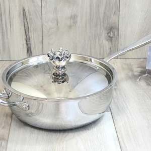 May include: A stainless steel saucepan with a glass lid and a decorative floral-shaped handle. The pan has a long handle and a smaller handle on the opposite side. A tag with text is visible.