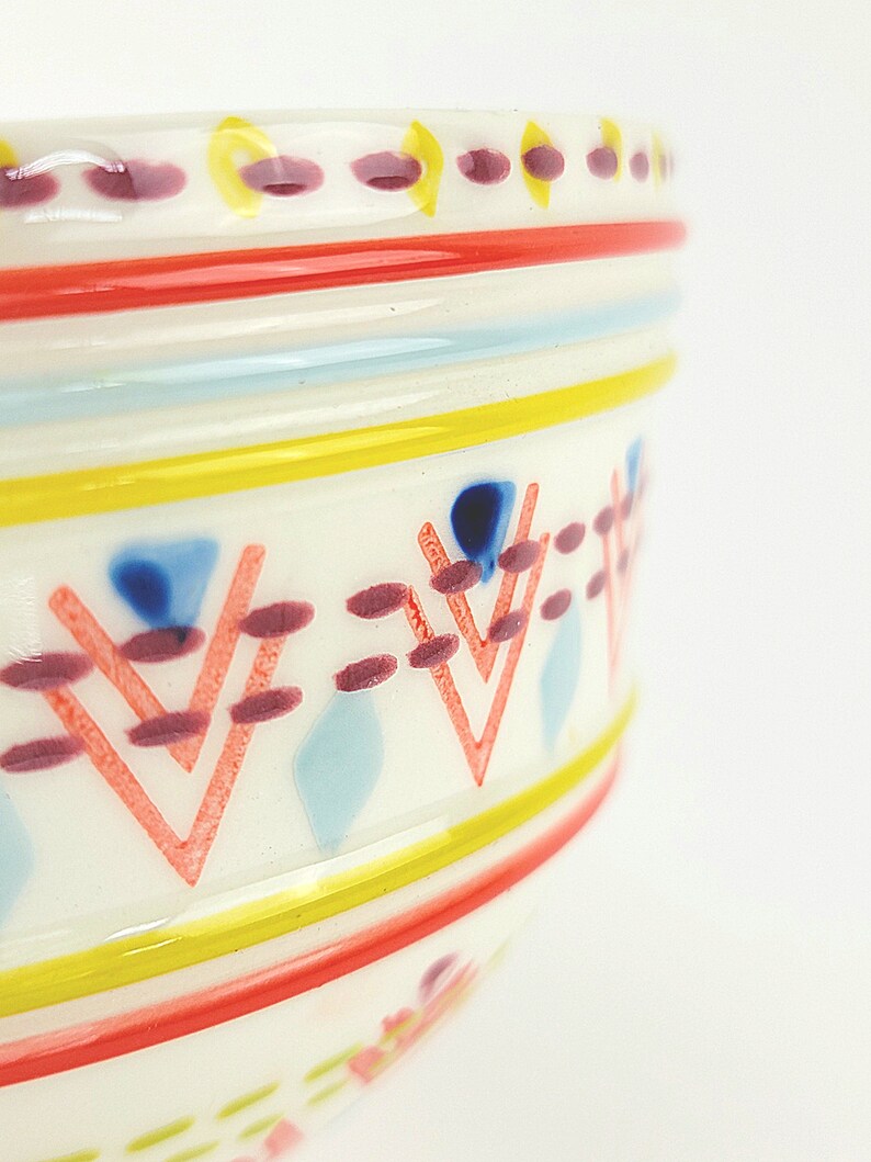 May include: A white ceramic bowl with a colourful geometric pattern. The pattern includes red, yellow, blue and pink stripes and triangles.