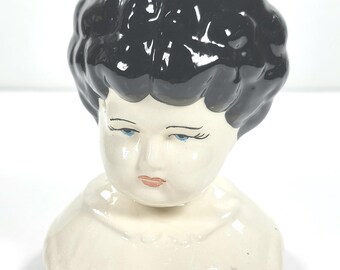 Może przedstawiać: Vintage ceramiczna głowa lalki z czarnymi, błyszczącymi włosami. Głowa lalki ma niebieskie oczy, delikatne rzęsy i małe, czerwone usta. Porcelanowa głowa jest w kolorze ecru i ma mały kołnierz. Dwa otwory są widoczne na dole szyi.