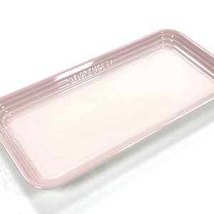May include: A rectangular light pink serving platter with a glossy finish and raised edges. The rim features a subtle three-ring embossed design. The brand name LE CREUSET is embossed in capital letters along one of the long sides of the platter.