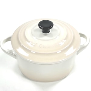 Puede incluir: Cazuela de hierro fundido esmaltado color crema con un pomo negro en la tapa. La olla tiene dos asas de bucle y el nombre de la marca está grabado en la tapa. Un artículo de cocina clásico.