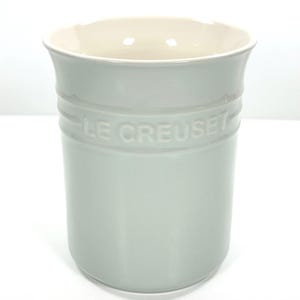 May include: A light green Le Creuset utensil holder with a cream-colored interior. The cylindrical holder features the brand name embossed around the middle. The kitchenware item is made of ceramic and has a smooth, glossy finish.