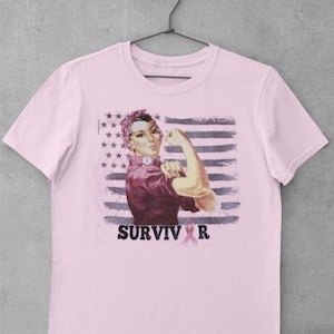 Puede incluir: Camiseta rosa claro con un diseño gráfico de una mujer flexionando su brazo, sobre un fondo de bandera americana. La palabra "SURVIVOR" está escrita en negro, con una cinta rosa.