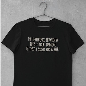 Könnte beinhalten: Schwarzes T-Shirt mit dem Text "THE DIFFERENCE BETWEEN A BEER & YOUR OPINION, IS THAT I ASKED FOR A BEER." Der weiße Text ist in einem handschriftlichen Stil gehalten. Das T-Shirt hängt an einem Metallbügel.