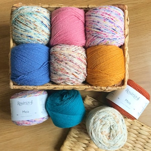 K&ouml;nnte beinhalten: Ein Weidenkorb gef&uuml;llt mit neun Garnkn&auml;ueln in verschiedenen Farben, darunter Rosa, Blau und gesprenkeltes Wei&szlig;. Mehrere zus&auml;tzliche Garnkn&auml;uel sind darunter zu sehen, mit Etiketten mit der Aufschrift "Rosarios 4 Meia Classic Collection."