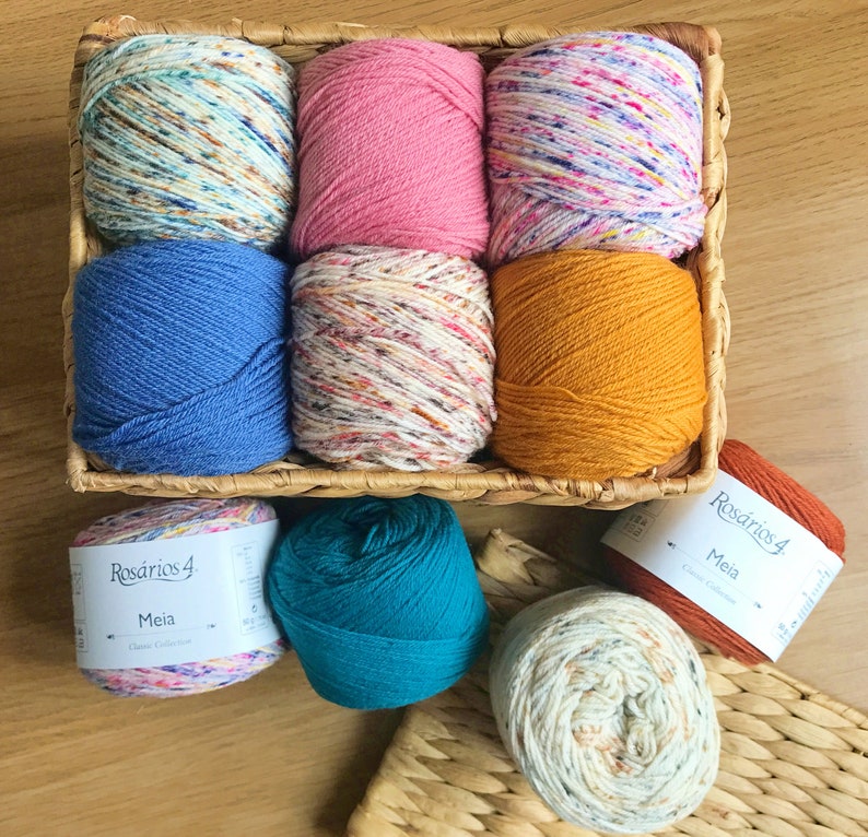 K&ouml;nnte beinhalten: Ein Weidenkorb gef&uuml;llt mit neun Garnkn&auml;ueln in verschiedenen Farben, darunter Rosa, Blau und gesprenkeltes Wei&szlig;. Mehrere zus&auml;tzliche Garnkn&auml;uel sind darunter zu sehen, mit Etiketten mit der Aufschrift "Rosarios 4 Meia Classic Collection."