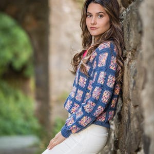K&ouml;nnte beinhalten: Ein blauer H&auml;kelpullover mit einem bunten Blumenmuster wird mit einer wei&szlig;en Hose getragen. Der Pullover hat ein quadratisches Muster mit einem durchgehenden blauen Rand. Der Hintergrund zeigt eine Steinmauer und einen verschwommenen gr&uuml;nen Bogen.