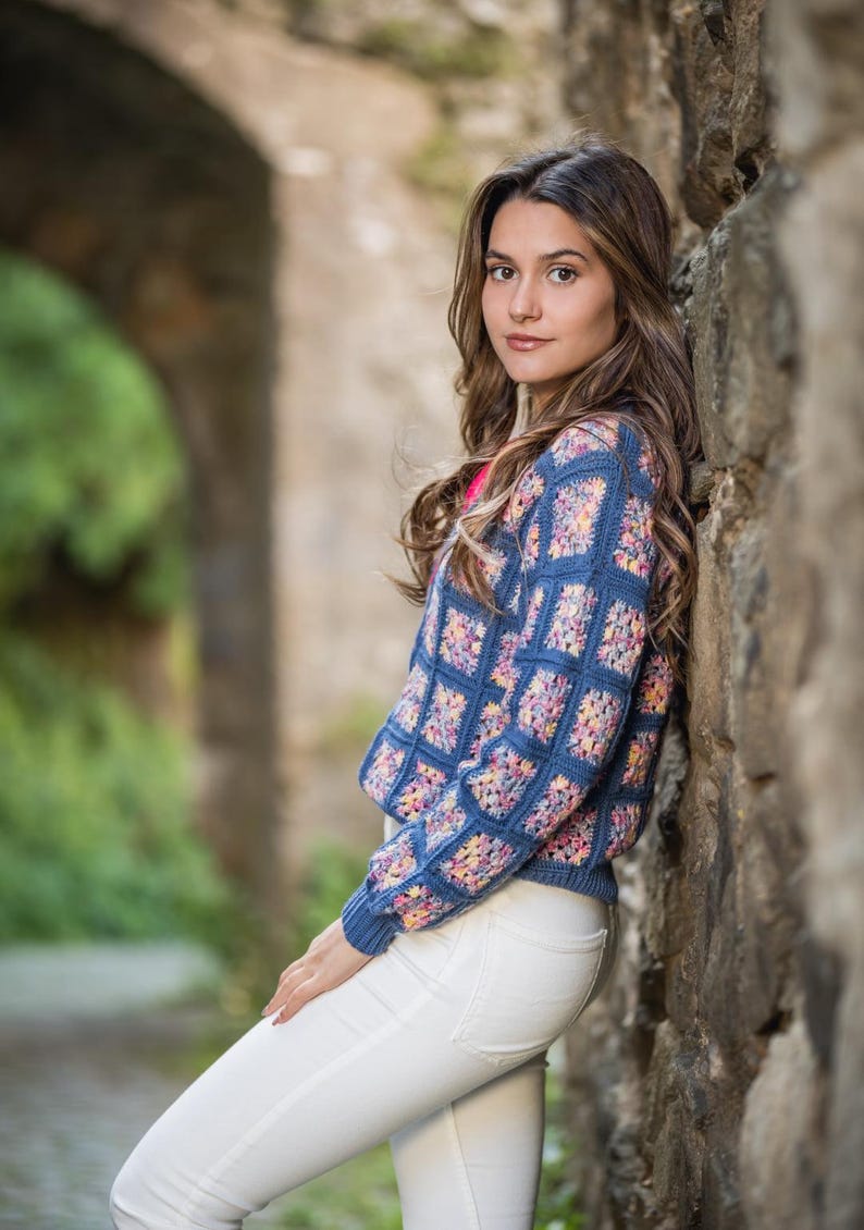K&ouml;nnte beinhalten: Ein blauer H&auml;kelpullover mit einem bunten Blumenmuster wird mit einer wei&szlig;en Hose getragen. Der Pullover hat ein quadratisches Muster mit einem durchgehenden blauen Rand. Der Hintergrund zeigt eine Steinmauer und einen verschwommenen gr&uuml;nen Bogen.