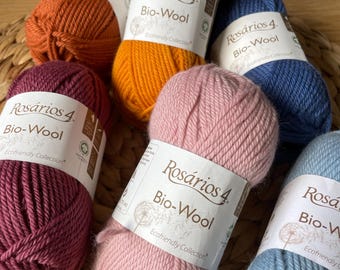 Könnte beinhalten: Sechs Knäuel Rosários 4 Bio-Wool Garn in verschiedenen Farben, darunter tiefes Burgunderrot, gebranntes Orange, Senfgelb, Marineblau, zartes Rosa und Hellblau. Jedes Knäuel hat ein weißes Papieretikett mit dem Markennamen und Produkttext.