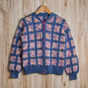 K&ouml;nnte beinhalten: Blaue H&auml;keljacke mit pinkfarbenem Rei&szlig;verschluss und quadratischem Muster. Die Jacke ist aus blauem Garn mit bunten Quadraten in Gelb, Rosa und Lila gefertigt. Die Jacke h&auml;ngt an einem Holzb&uuml;gel.