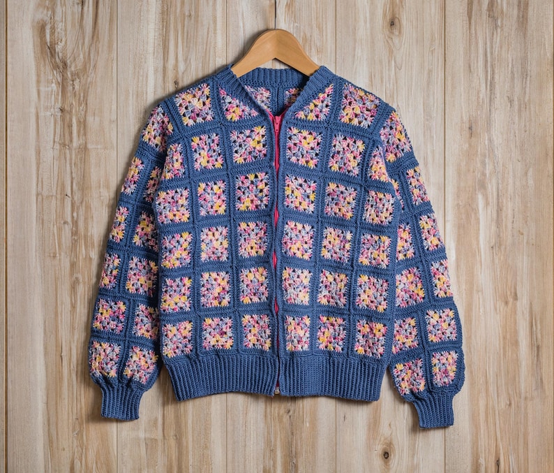 K&ouml;nnte beinhalten: Blaue H&auml;keljacke mit pinkfarbenem Rei&szlig;verschluss und quadratischem Muster. Die Jacke ist aus blauem Garn mit bunten Quadraten in Gelb, Rosa und Lila gefertigt. Die Jacke h&auml;ngt an einem Holzb&uuml;gel.