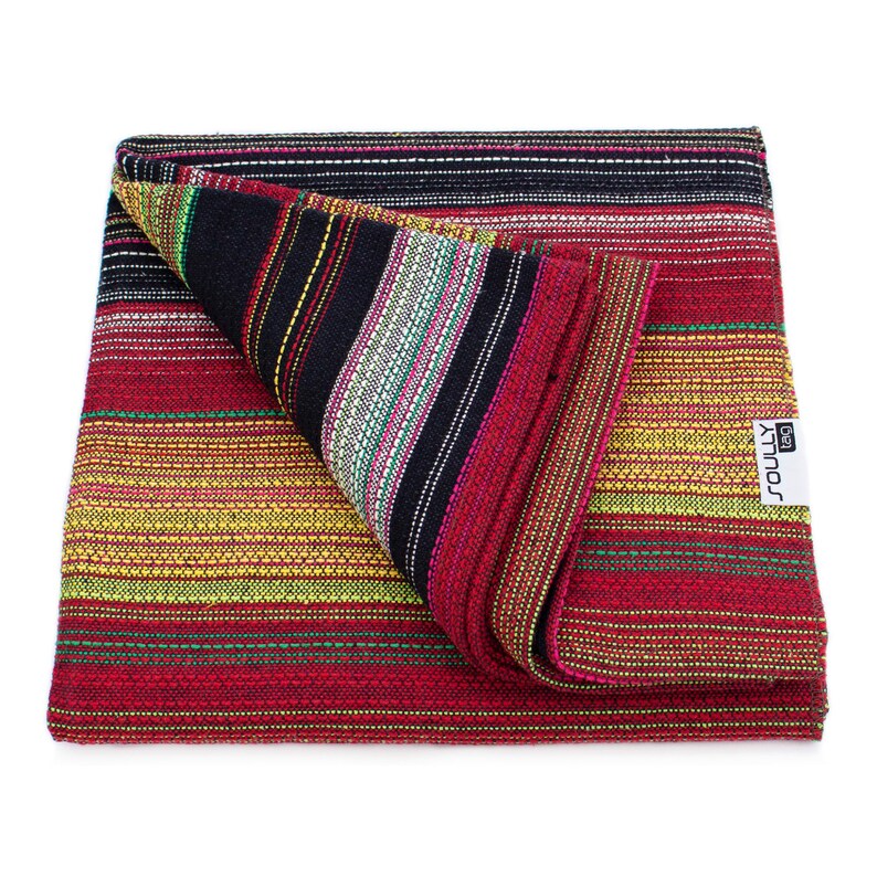 May include: A folded woven textile featuring a vibrant striped pattern in shades of red, yellow, black, and green. The fabric has a textured appearance with horizontal lines and a white rectangular tag with the text SOULLY tag attached to the side.