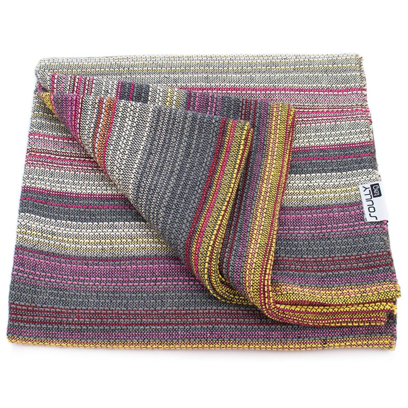 May include: A folded woven blanket featuring a multi-colored striped pattern in shades of gray, magenta, yellow, and white. The fabric has a textured, interlaced weave throughout. A white rectangular tag with the black text SOULLY tag is attached.