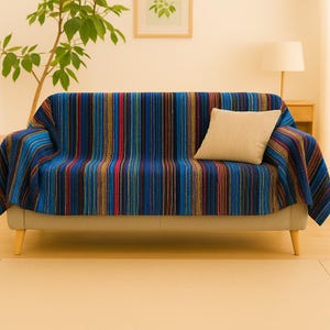 May include: A sofa covered with a multi-colored striped throw blanket featuring vertical lines in shades of blue, red, yellow, and black. A single solid beige square throw pillow rests on the right side of the seat cushion.