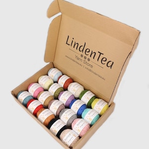 Pode incluir: Uma caixa de cartão aberta cheia de novelos de lã coloridos. A caixa contém fios de várias cores, incluindo azul, rosa, amarelo e verde. A caixa tem o texto "LindenTea Yarn Store" impresso.