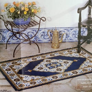 Può includere: Un tappeto rettangolare blu navy con un motivo floreale bianco, oro e blu. Il tappeto è posizionato su un pavimento in pietra, con un supporto decorativo in metallo con un vaso blu di rose gialle e una brocca dorata.
