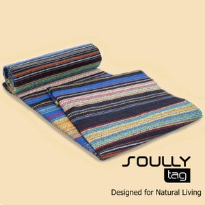 May include: A woven fabric runner featuring a multi-colored striped pattern with bands of black, blue, yellow, and white. The textile is partially rolled and partially laid flat. The logo SOULLY tag and the text Designed for Natural Living are visible.