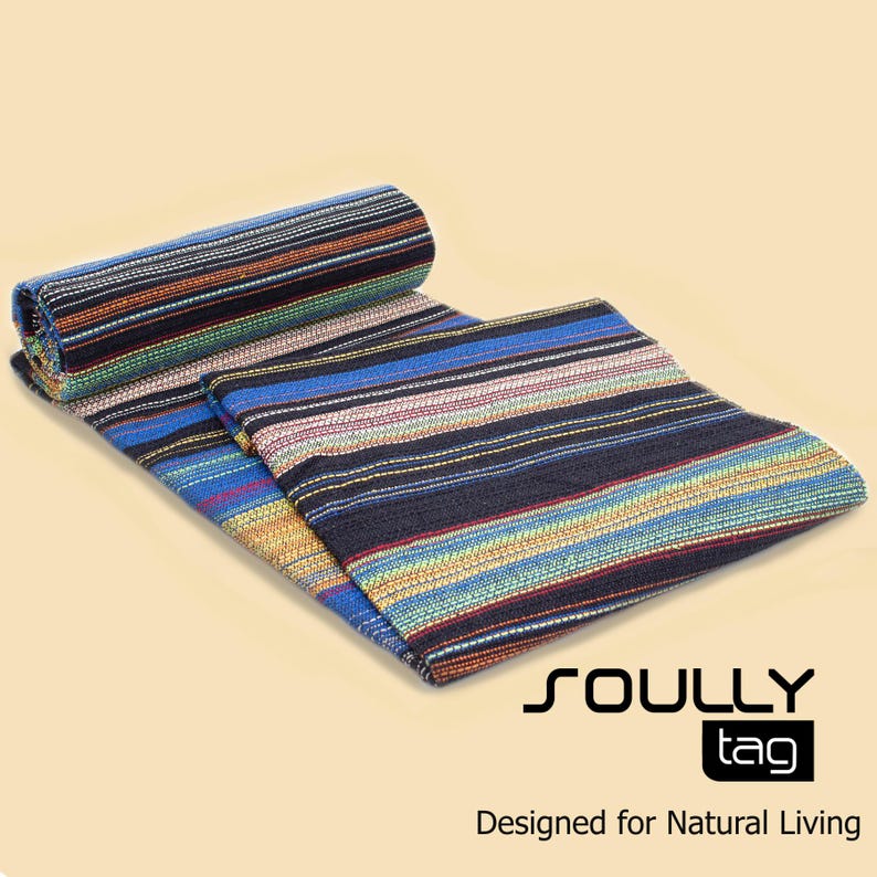May include: A woven fabric runner featuring a multi-colored striped pattern with bands of black, blue, yellow, and white. The textile is partially rolled and partially laid flat. The logo SOULLY tag and the text Designed for Natural Living are visible.