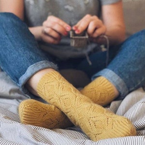 K&ouml;nnte beinhalten: Senfgelbe Strickstr&uuml;mpfe mit detailliertem Lochmuster. Die Socken werden mit einer blauen Jeans getragen. Eine Person strickt einen kleinen Gegenstand, mit den H&auml;nden und dem Strickprojekt im Hintergrund.