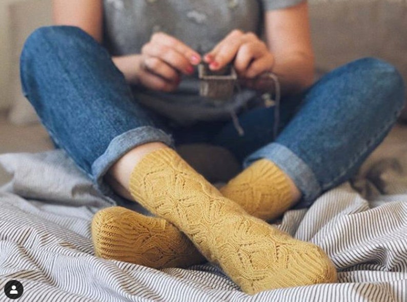 K&ouml;nnte beinhalten: Senfgelbe Strickstr&uuml;mpfe mit detailliertem Lochmuster. Die Socken werden mit einer blauen Jeans getragen. Eine Person strickt einen kleinen Gegenstand, mit den H&auml;nden und dem Strickprojekt im Hintergrund.