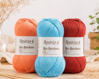 以下が含まれることがあります： オレンジ、ブライトブルー、ダークレッドのRosários 4 Bio Bamboo毛糸玉3個。各毛糸玉には、ブランド名、製品名、Ecofriendly Collectionという文字が印刷された白い紙のラベルが付いています。手前には木製の編み針が2本置かれています。