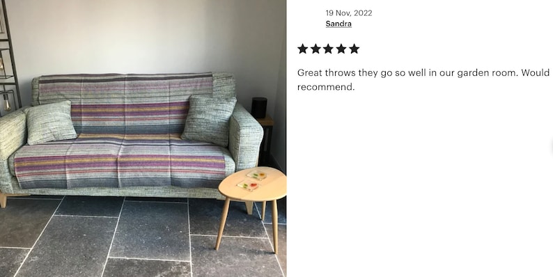 May include: A woven throw blanket with horizontal stripes in shades of purple, yellow, grey, and blue draped over a light green upholstered sofa. Two matching throw pillows are placed on the sofa, which features light-colored wooden legs.