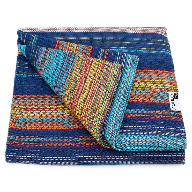 May include: A folded woven blanket featuring a vibrant striped pattern in shades of deep blue, turquoise, yellow, orange, and red. The fabric has a textured appearance with horizontal lines and a white rectangular tag labeled SOULLY tag.