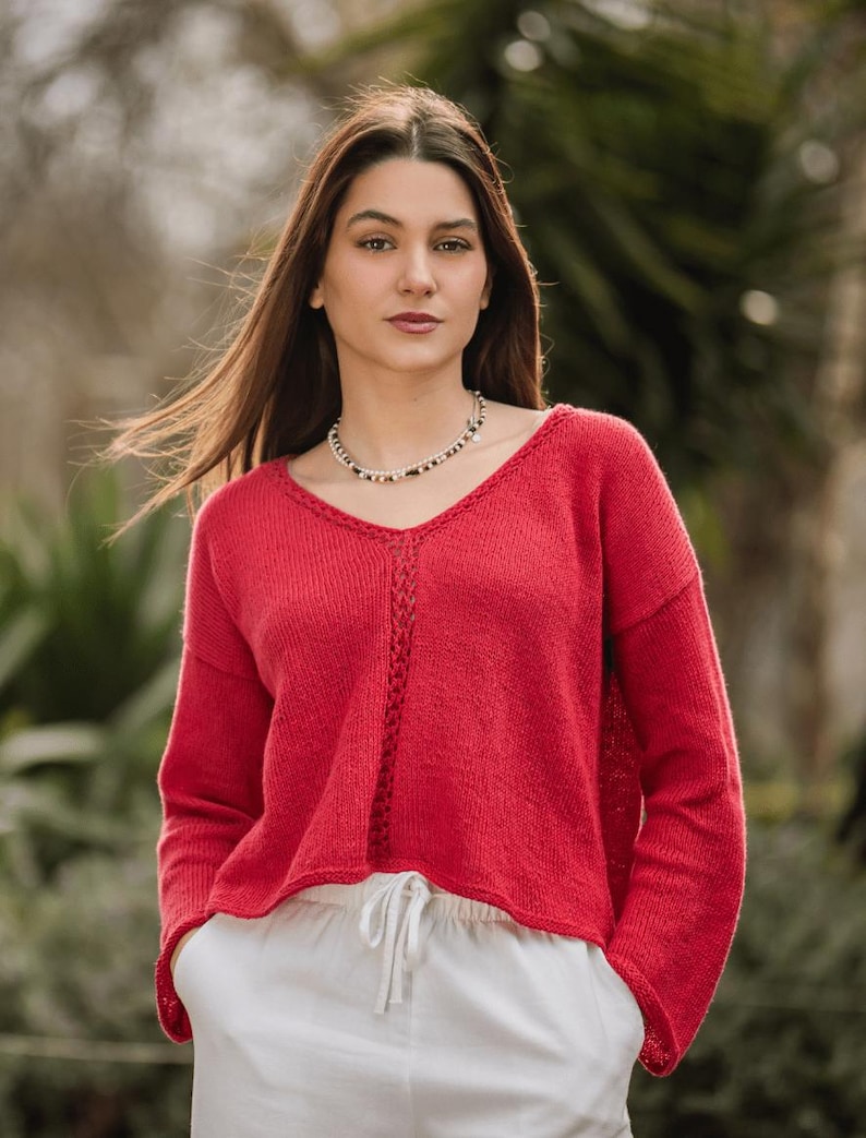 Puede incluir: Un su&eacute;ter rojo vibrante de manga larga con cuello en V y un detalle central de punto trenzado vertical, combinado con pantalones blancos con cintura de cord&oacute;n. El su&eacute;ter tiene un corte relajado con hombros ca&iacute;dos y pu&ntilde;os acanalados.