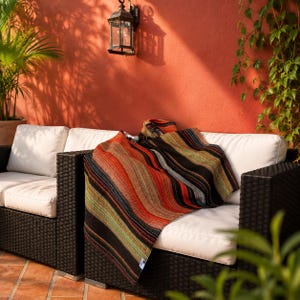 May include: A woven throw blanket featuring a multi-colored striped pattern in shades of black, orange, red, and green. The blanket is draped over a black wicker outdoor sofa with white cushions, showcasing a textured fabric with vertical lines.