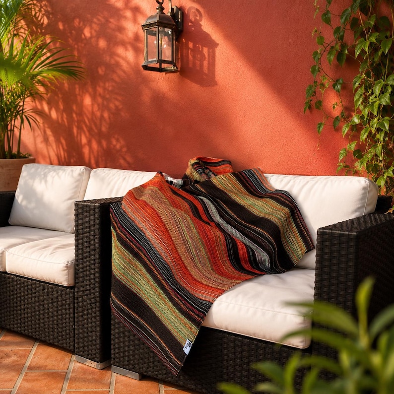 May include: A woven throw blanket featuring a multi-colored striped pattern in shades of black, orange, red, and green. The blanket is draped over a black wicker outdoor sofa with white cushions, showcasing a textured fabric with vertical lines.