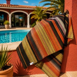 May include: A rectangular woven textile featuring a vertical striped pattern in shades of black, burnt orange, olive green, and cream. The fabric has a textured appearance with a small white rectangular label attached to the lower corner reading Soulia.