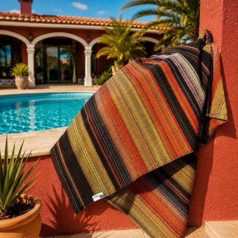 May include: A rectangular woven textile featuring a vertical striped pattern in shades of black, burnt orange, olive green, and cream. The fabric has a textured appearance with a small white rectangular label attached to the lower corner reading Soulia.