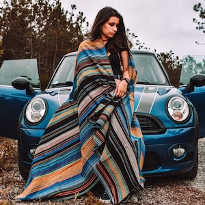 May include: A large, rectangular woven blanket featuring a vibrant, multi-colored striped pattern in shades of blue, orange, black, and white. The fabric displays a textured, dense weave with horizontal lines running across the entire length of the textile.