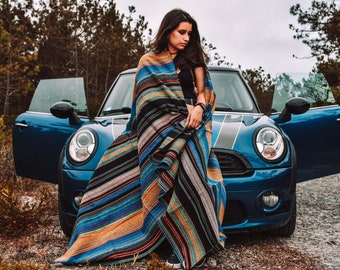 May include: A large, rectangular woven blanket featuring a vibrant, multi-colored striped pattern in shades of blue, orange, black, and white. The fabric displays a textured, dense weave with horizontal lines running across the entire length of the textile.