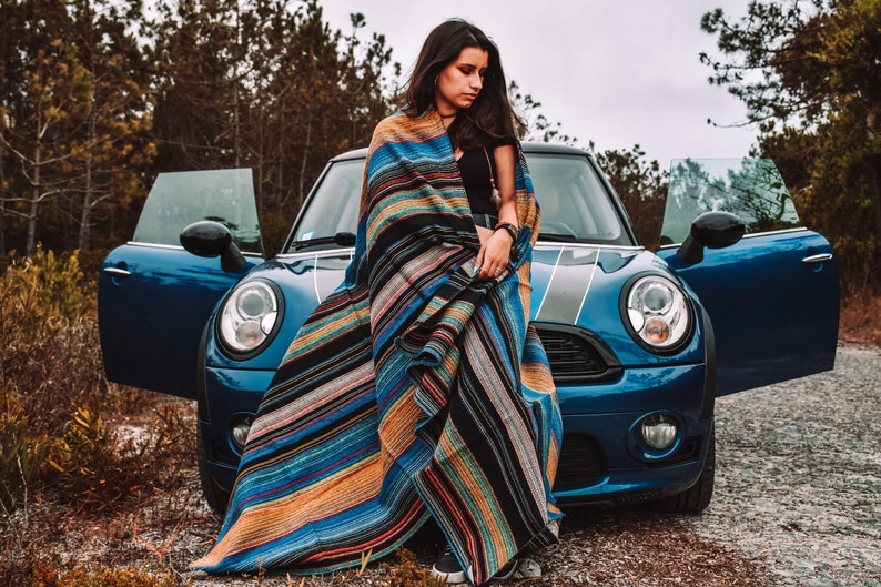 May include: A large, rectangular woven blanket featuring a vibrant, multi-colored striped pattern in shades of blue, orange, black, and white. The fabric displays a textured, dense weave with horizontal lines running across the entire length of the textile.