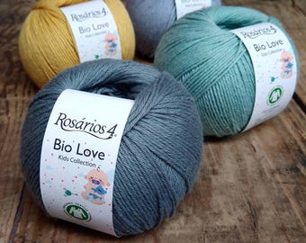 Puede incluir: Cuatro madejas de hilo en tonos amarillo, gris y verde azulado. Cada madeja tiene una etiqueta que dice "Rosários 4 Bio Love Kids Collection". El hilo está dispuesto sobre una superficie de madera.