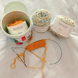 K&ouml;nnte beinhalten: Draufsicht auf Strickzubeh&ouml;r. Es gibt drei Kn&auml;uel Garn, zwei mit einem gesprenkelten Muster und eines in Orange. Ein teilweise fertiges orangefarbenes Strickmuster liegt auf Nadeln. Das Garn ist mit "Ros&aacute;rios 4 Meia" beschriftet.